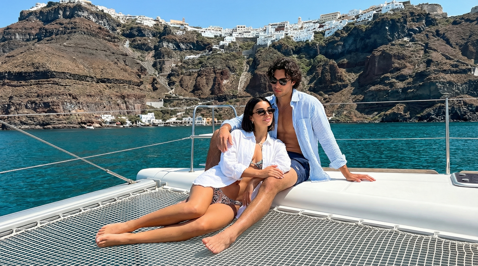 Couple on catamaran sailing the Santorini caldera with Oia cliff face in background